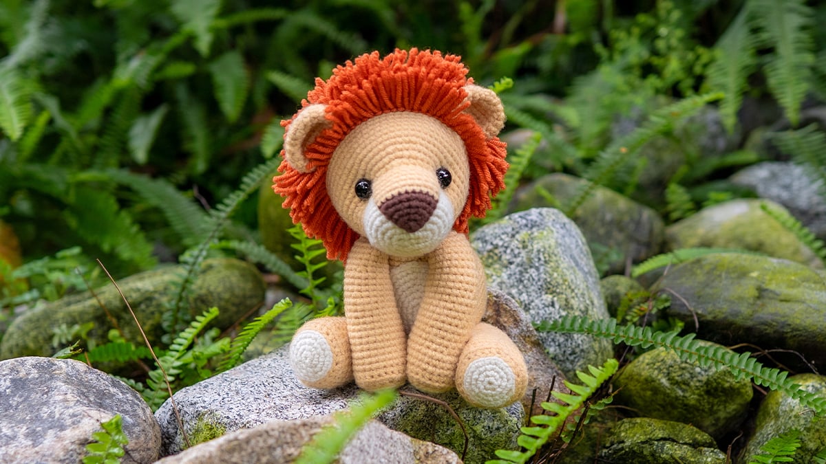 Yellow crocheted amigurumi lion with a fluffy orange mane and black safety eyes sitting on a stone in a jungle setting, made with a free lion crochet pattern.”