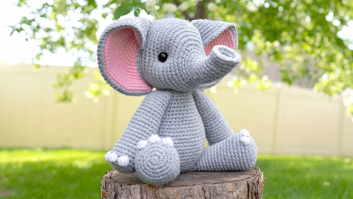 Light gray crocheted amigurumi elephant with large pink-lined ears, black safety eyes, and a curved trunk, sitting on a tree stump outdoors, made from a free elephant crochet pattern.