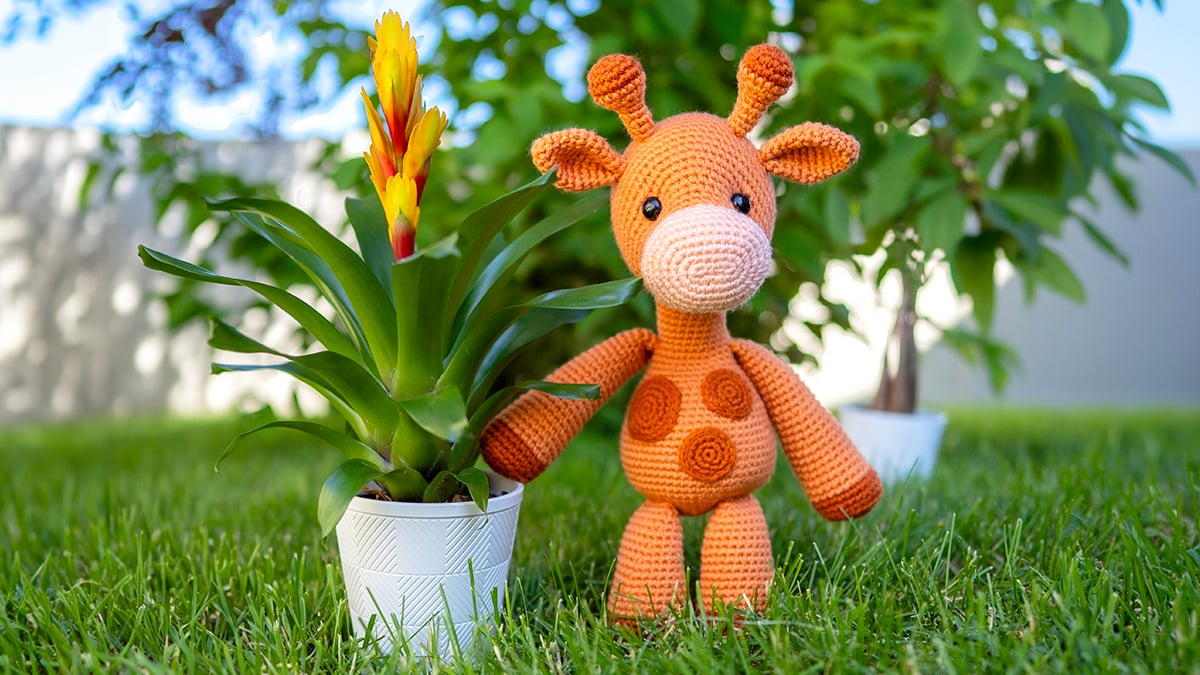 Orange crocheted amigurumi giraffe with long arms and dark orange spots, standing on grass in nature, made with a free giraffe crochet pattern.