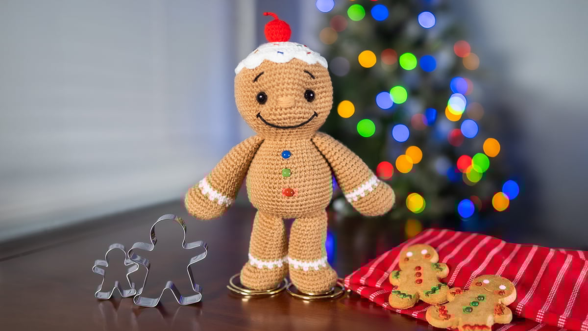 Crocheted amigurumi gingerbread man with icing details, colorful buttons, and a cherry on top standing on a wooden table beside metal cookie cutters and gingerbread cookies, with a blurred Christmas tree in the background, made with a free gingerbread crochet pattern.