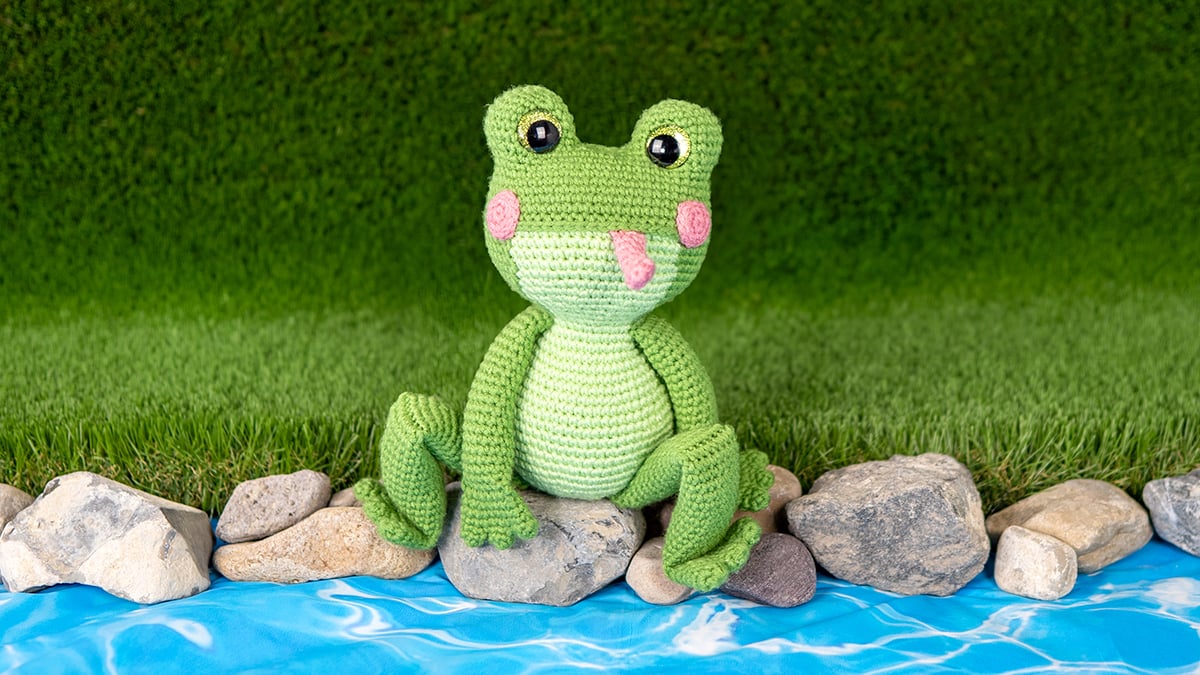 Green crocheted amigurumi frog with big sparkly yellow safety eyes, sitting on rocks by a river, made with a free frog crochet pattern.