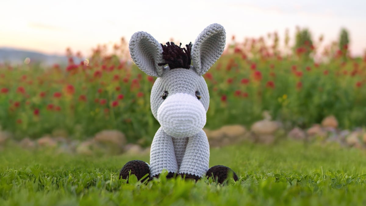 Gray crocheted amigurumi donkey with long ears, a white muzzle, and a black mane, sitting on grass with red roses blurred in the background, made with a free donkey crochet pattern.