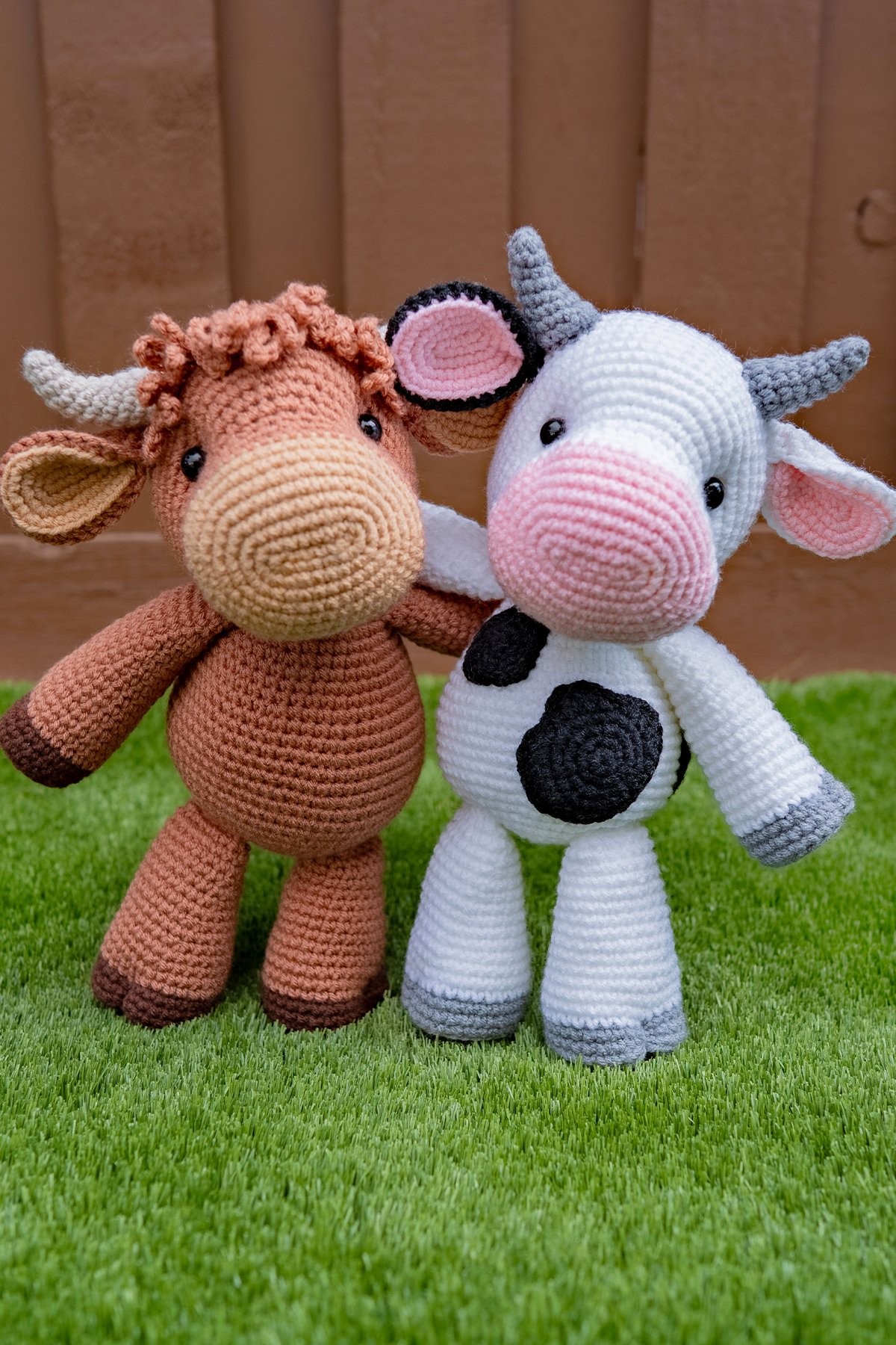 Holly the Highland Cow and Caroline the Cow crocheted amigurumi standing together on grass with their arms around each other