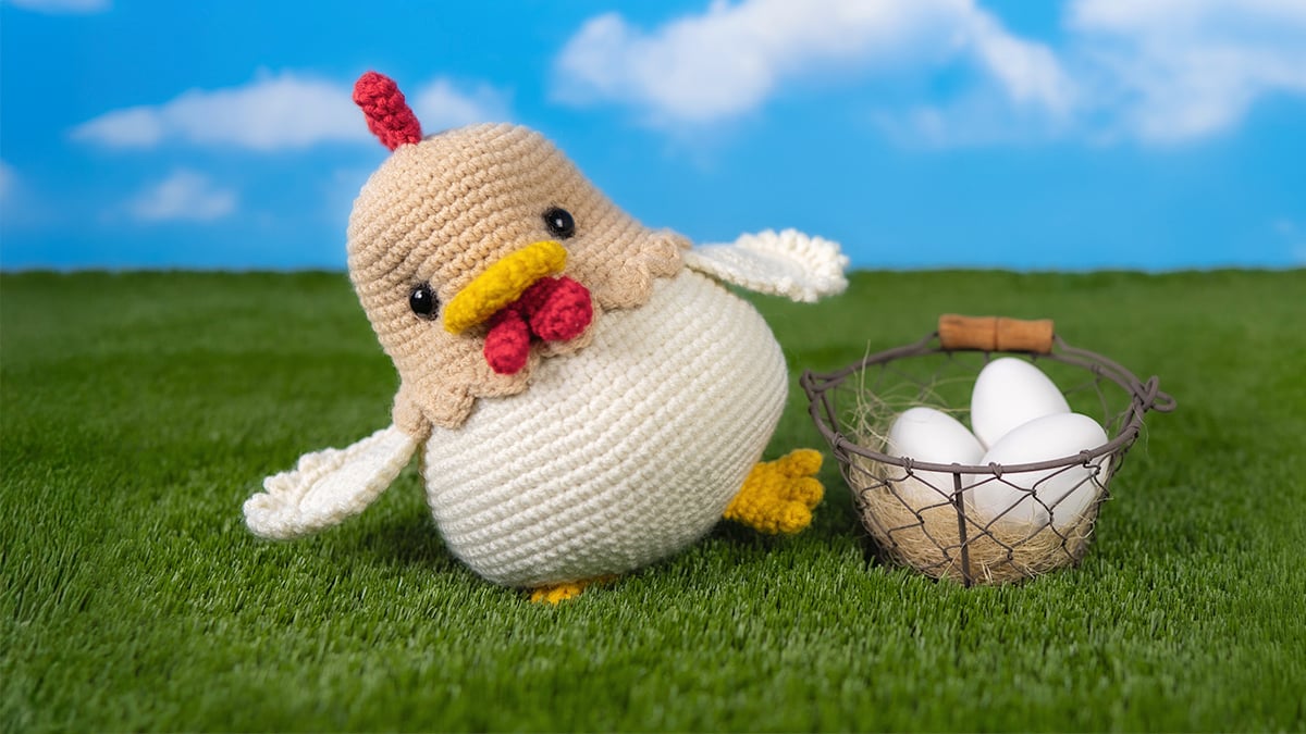 Handmade amigurumi chicken crocheted in cream and tan yarn, standing on green grass beside a wire basket filled with white eggs, photographed against a blue sky backdrop.