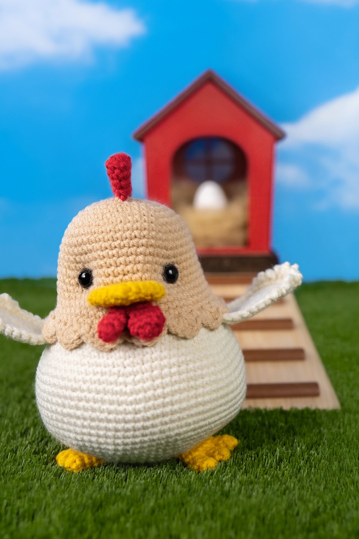 Gratis Kiki Kip Haakpatroon 1 Handmade amigurumi chicken crocheted in cream and tan yarn, standing on green grass with wings slightly raised, with a red chicken coop and a white egg visible in the background against a blue sky.