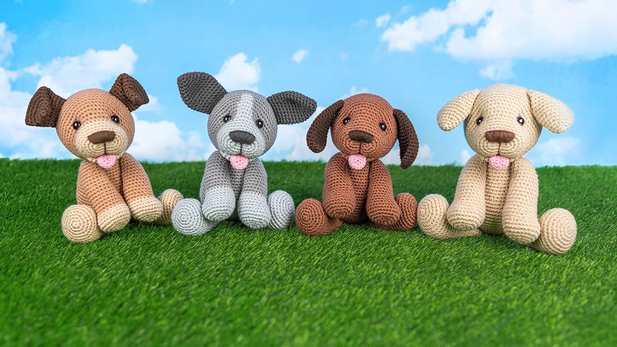 Group of crocheted amigurumi dogs in shades of brown, tan, and cream displayed together on green grass, made from a free dog crochet pattern.