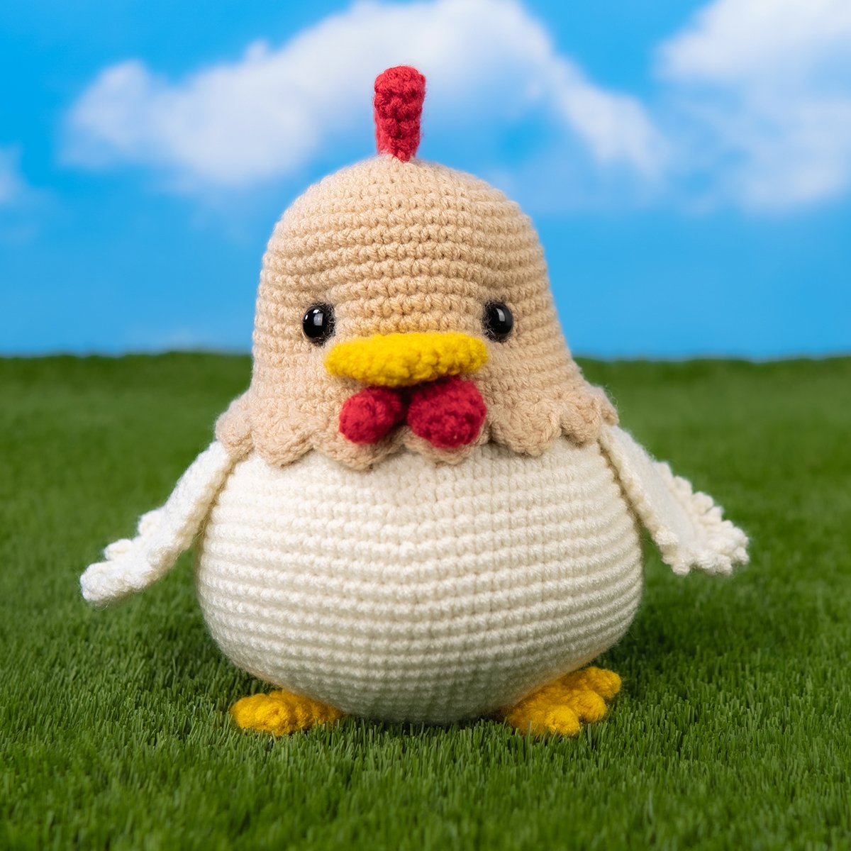 Gratis Kiki Kip Haakpatroon 12 Free chicken crochet pattern featuring a round amigurumi chicken with a cream body, beige head, red comb and wattle, yellow beak and feet, standing on grass against a blue sky background.