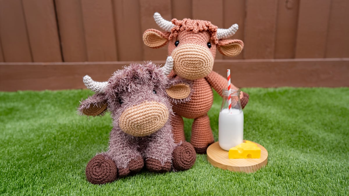 Two highland cow stuffed animals—one crocheted with fluffy eyelash yarn and one crocheted with standard acrylic—sitting together on green grass beside a glass milk bottle and cheese.