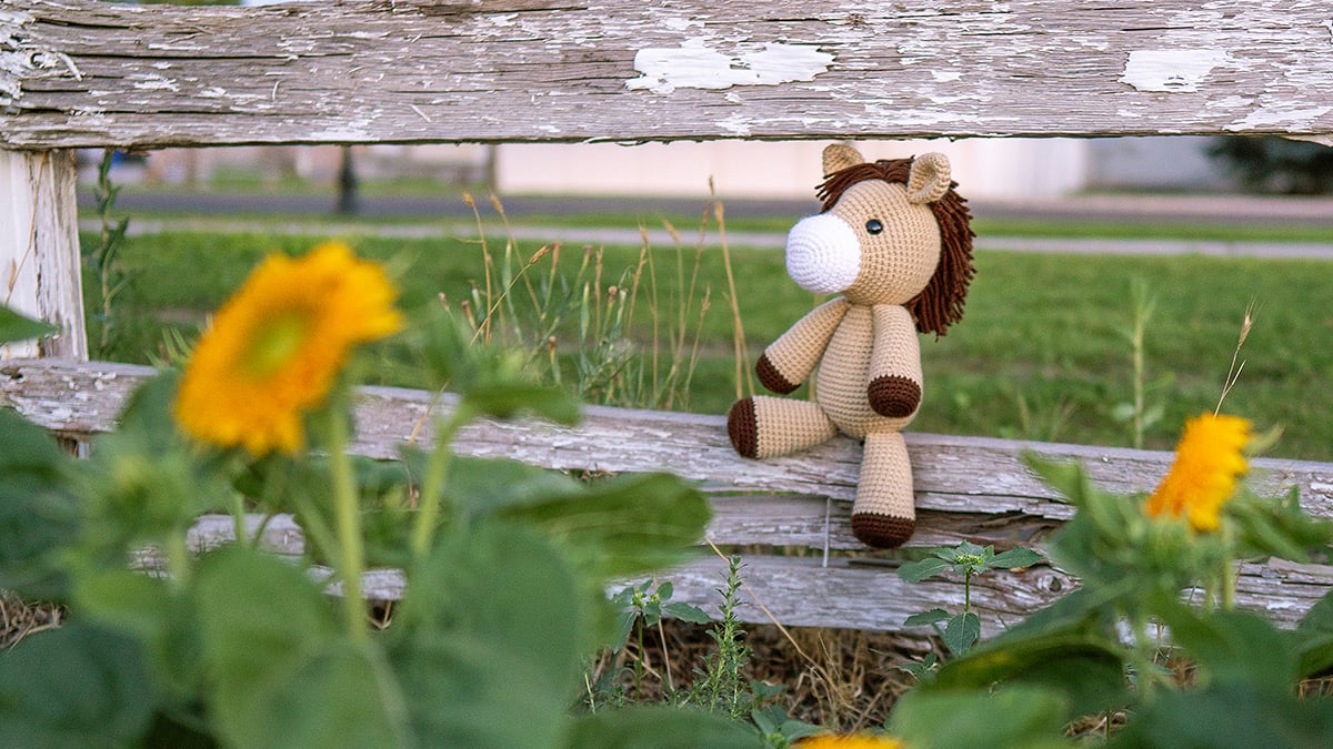 Light brown crocheted amigurumi horse with cream muzzle and dark brown mane, sitting on a rustic wood fence surrounded by grass and sunflowers, made with a free horse crochet pattern.