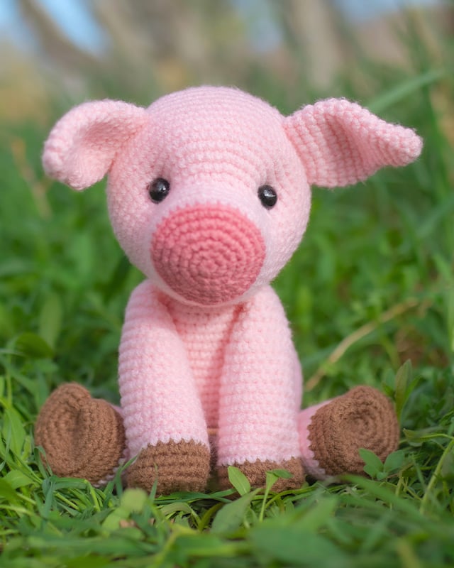 Free pig crochet pattern featuring a pink amigurumi pig with brown hooves sitting on grass.