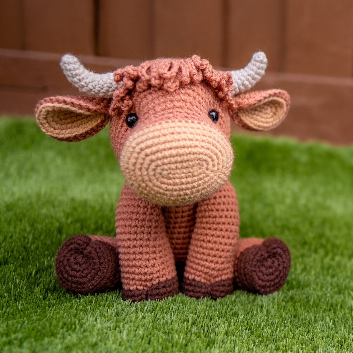 Handmade crocheted highland cow stuffed animal made from a free amigurumi pattern, with loopy bangs, curved horns and safety eyes, photographed on green grass.