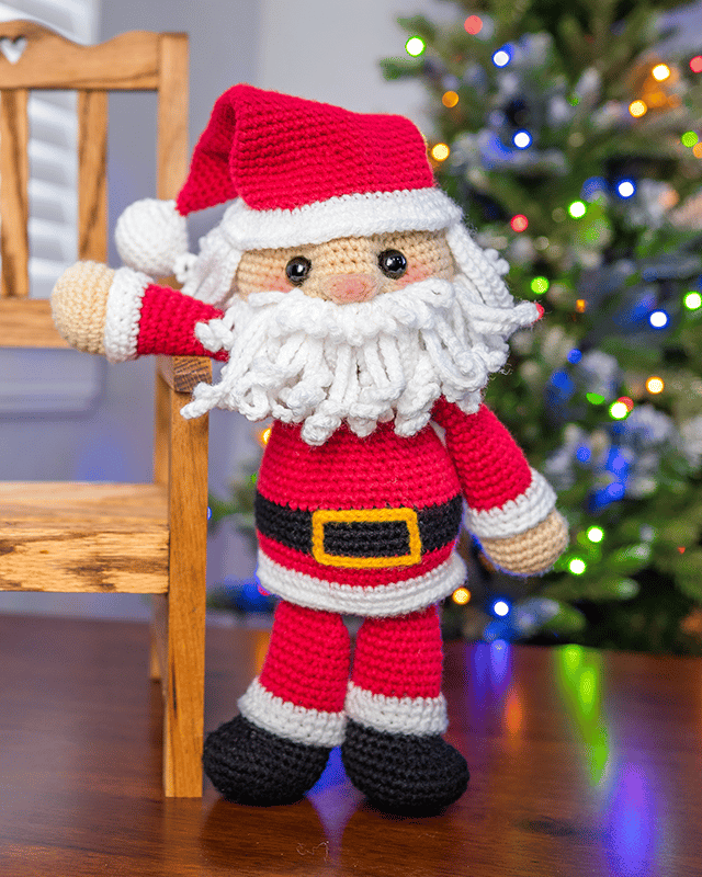 Crocheted Santa amigurumi doll standing beside a wooden chair with a lit Christmas tree in the background.